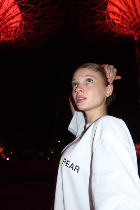 A young woman with light hair poses against a dark background with red string lights overhead, wearing a white jacket with the word "PEAR" printed on it.