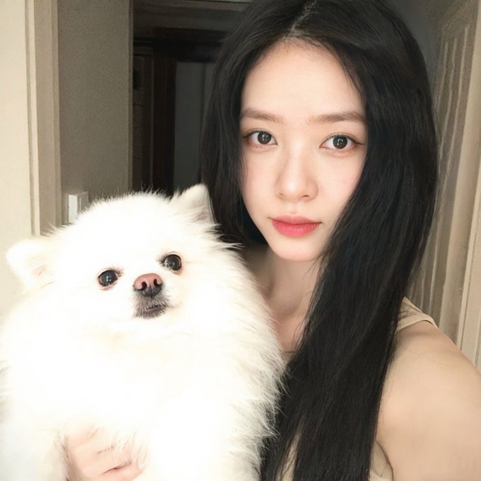 A selfie of a young woman with long black hair and red lipstick holding a fluffy white Pomeranian dog.
