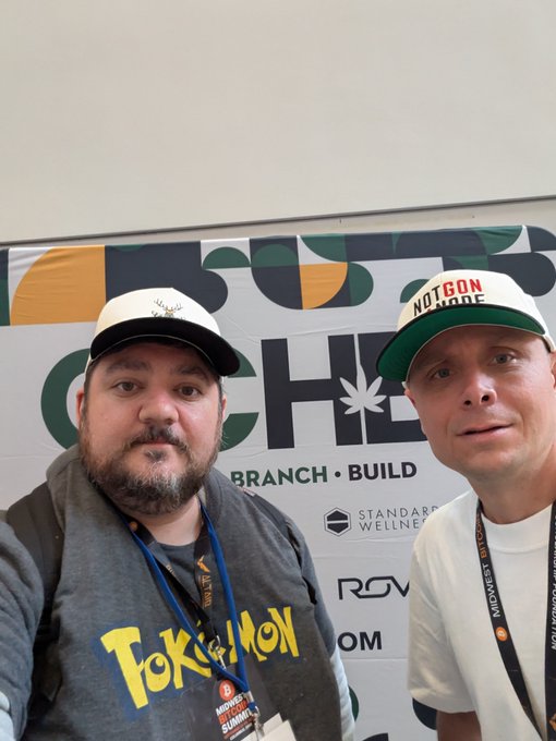 Two men posing for a selfie in front of a 'NOTGON MODE' banner; one wears a 'Pokémon' shirt and the other wears a white cap with the same event logo, both with lanyards.