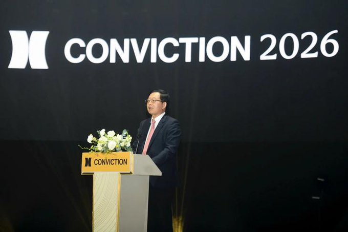 A speaker in a dark suit and pink tie stands at a podium labeled "CONVICTION" with a matching large backdrop reading "CONVICTION 2026," illustrating the Vietnamese message about digital assets evolving beyond speculative crypto into a broader strategic technology story combining blockchain and AI.