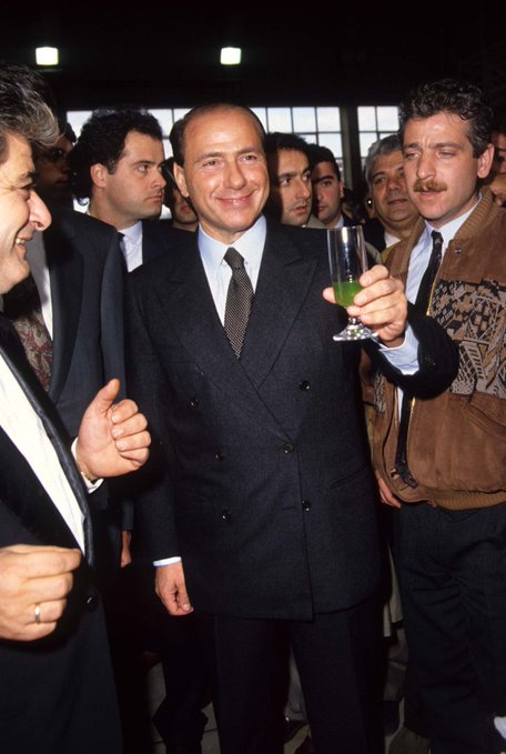 A man in a dark double-breasted suit and tie smiles while holding up a glass of green liquid at what appears to be a social gathering, surrounded by other attendees.