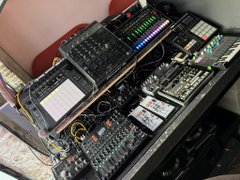 A close-up view of professional audio/sound mixing equipment with multiple control panels, knobs, faders, and a small screen, with cables connected.