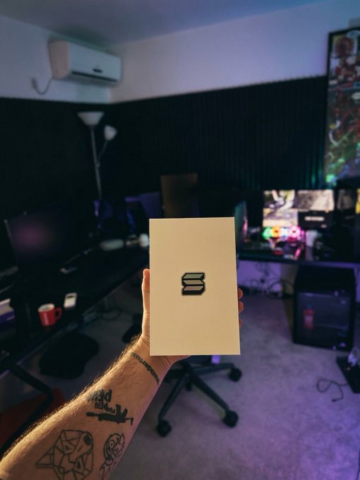 A tattooed arm holds up a beige card with a black geometric 'S' logo in a dimly lit gaming/streaming room setup.