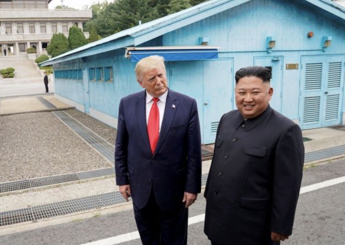 Donald Trump in a dark suit and red tie stands next to Kim Jong-un in a black Mao-style suit in front of a blue building at the Korean Demilitarized Zone.
