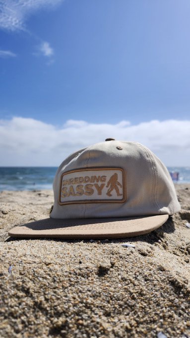 A beige and brown baseball cap with "SHREDDING SASSY" text and a Bigfoot patch resting on sandy beach.