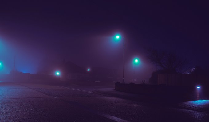 A foggy nighttime street scene with green-tinted streetlights glowing through dense mist, casting a moody purple-blue atmosphere over an empty road and silhouetted buildings.
