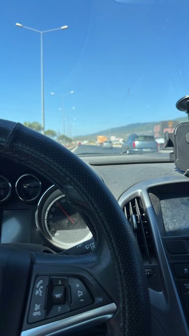 A view from the driver's seat of a car on a sunny day, showing the steering wheel, dashboard speedometer, and a highway ahead with other vehicles.