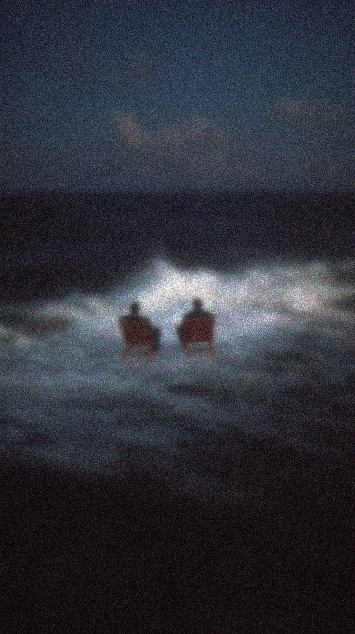 A dark, atmospheric photo showing two silhouetted figures standing in ocean water with waves breaking behind them and a small patch of lighter sky visible.