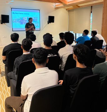 The image shows a group of people sitting in chairs, watching a presentation or lecture being given by someone standing behind a screen. The audience appears engaged and attentive as they listen to the speaker's content. The room has a white wall with a projector screen mounted on it, which is likely used for presentations or discussions.