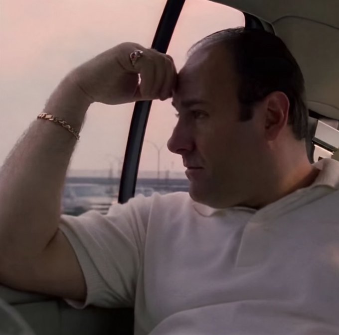 A side profile shot of a man sitting in a vehicle, resting his hand on his forehead while wearing a white t-shirt, a gold ring, and a gold bracelet.