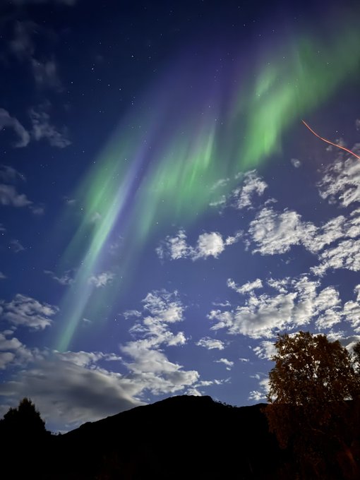 A night sky view showcasing vibrant green and purple aurora borealis stretching above scattered clouds and the dark silhouette of a mountain ridge.