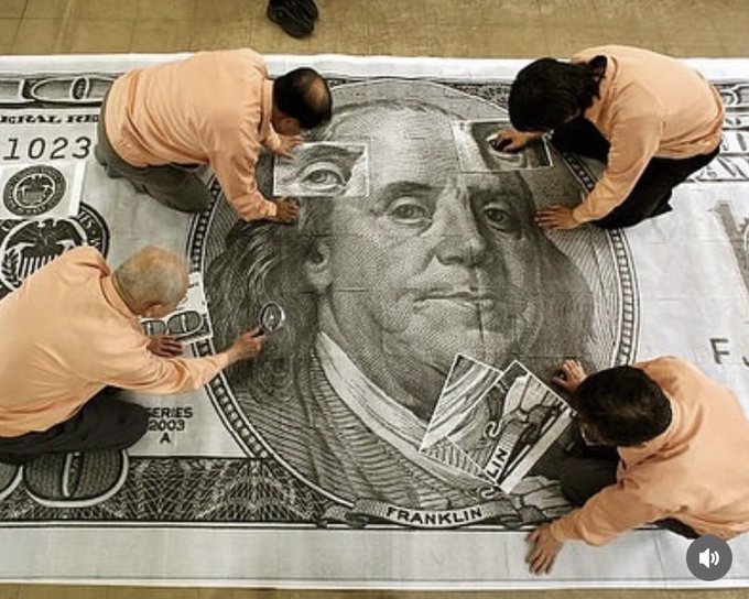 Four people in peach-colored shirts kneeling around an oversized replica of a US 100-dollar bill featuring Benjamin Franklin, working on details with magnifying glasses.