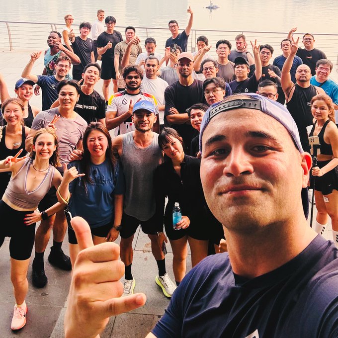 A large group of people in athletic wear pose together for a selfie near a waterfront, with a man in a backwards cap giving a 'shaka' hand gesture in the foreground.