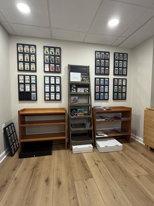 The image features a room with wooden floors, white walls, and shelves filled with cards. The room appears to be an office or storage area for collecting cards. There are multiple shelves holding various cards, including some that have been collected over time. A few boxes can also be seen in the room, possibly containing additional card collections or supplies. The overall atmosphere of the space suggests a dedicated area for card enthusiasts to store and display their valuable items.