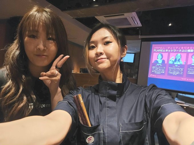 Two women pose for a selfie at what appears to be a tech or cryptocurrency event, with a presentation slide in the background reading "XRPのユーティリティを引き出す FLARE＆ネットワーク上に構築" (Unlocking the utility of XRP - Built on the FLARE & network), referencing the Flare Network and its connection to XRP, aligning with the post about the Flare×Tangem side event.