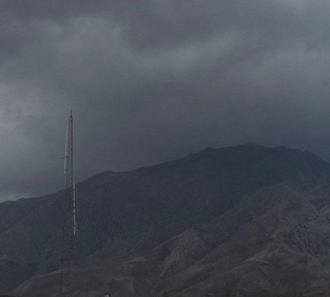 A tall communications tower stands on the left against a backdrop of dark, cloudy skies and rugged, barren mountain terrain.