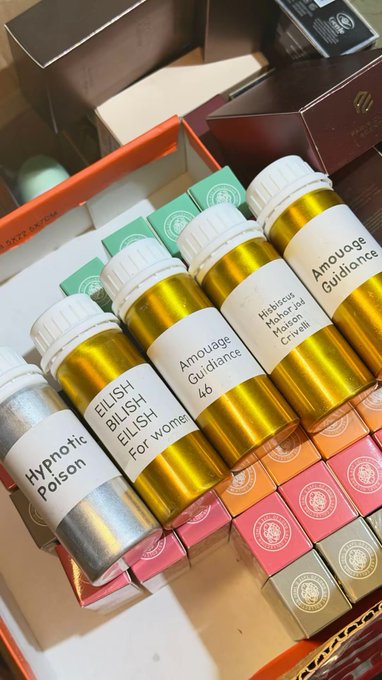 A collection of small bottles with whimsical labels including 'Hypnotic Poison', 'EILISH EILISH For wome', 'Amouage Guidance', and 'Nishane Mancera Crisvelli' displayed among gift boxes.