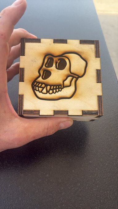 A hand holds up a small laser-cut wooden box featuring an engraved gorilla skull design on its lid, tied to a giveaway post promoting @ApeChainHUB and @BoredApeYC.