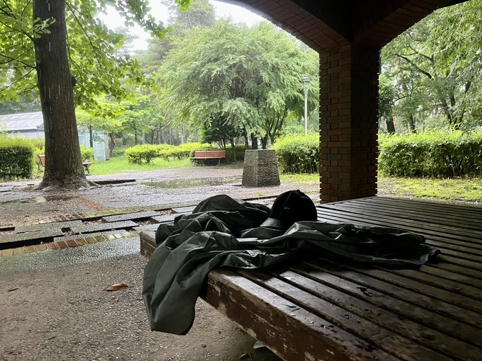 The image shows a wooden bench with a black jacket draped over one end, sitting under an archway or pavilion. The bench appears to be wet from rainwater, suggesting that it has been recently used by someone who was seeking shelter during a downpour. The surrounding area is filled with trees and greenery, creating a peaceful atmosphere. The post in the image reads "ファミマでポンチョ買った。ネカフェでシャワー借りてやる事行き詰まってるので12時間パックでシコります". This post is likely a reference to the bench's location, as it provides information about where the bench can be found. The wet bench and surrounding environment suggest that this scene takes place in an outdoor park or public space during rainy weather.