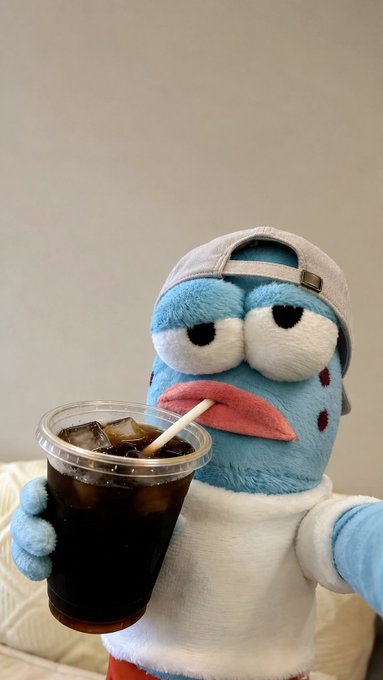A blue plush bird toy wearing a white cap and white shirt holds a plastic cup of iced dark soda with a straw.