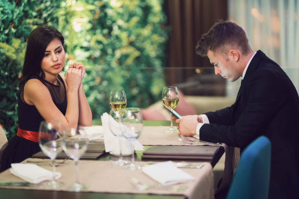 A tense scene at a restaurant with a woman in a black dress looking annoyed across the table at a man in a suit who is distracted by his phone, with wine glasses between them.
