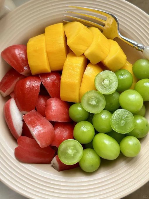 A cream-colored bowl filled with fresh fruit, including sliced red apples, yellow mango chunks, and green grapes, with a silver fork resting on the edge.