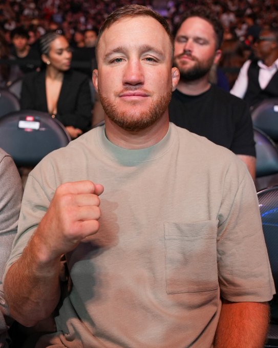 A red-bearded man in a beige t-shirt raises his fist at a crowded UFC event, seated among other spectators in arena chairs, fitting the social media caption promoting the live #UFC331 broadcast on ParamountPlus sponsored by Crypto.com.