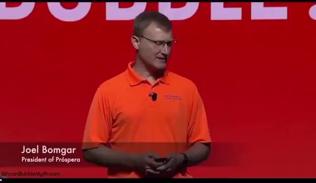 The image features a man wearing an orange shirt standing in front of a red background with white text that reads "Double Bubbles". The man appears to be giving a speech or presentation, possibly related to Bitcoin or cryptocurrency. The caption at the bottom of the image suggests that this event is being covered by someone named Joel Bombar on his blog.