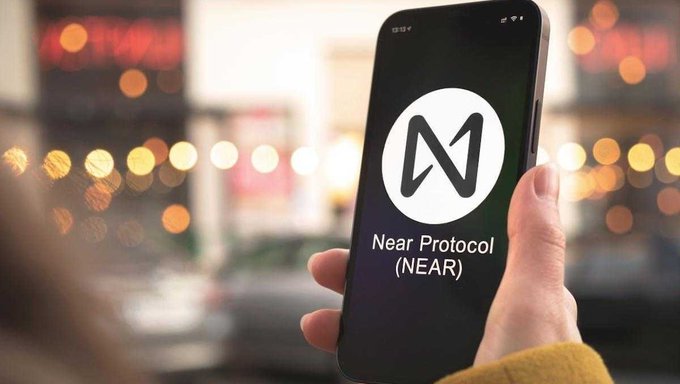 A hand holds a smartphone displaying the Near Protocol (NEAR) logo, with a blurred city street and bokeh lights in the background.