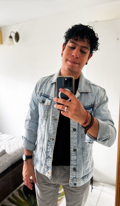 A mirror selfie of a young man with curly dark hair wearing a light blue denim jacket over a black shirt, taking a photo with a smartphone in a bedroom setting.