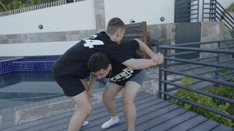 The image shows two young men playing a game of wrestling, with one man on top of the other as they engage in friendly competition. Both individuals are wearing black t-shirts that say "4". The background features a house and a pool, suggesting an outdoor setting where the wrestling match is taking place.