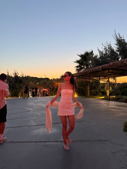 A woman in a short white strapless dress twirls playfully outdoors at sunset, wearing sunglasses and light-colored heels, with palm trees and a patio structure visible in the background.