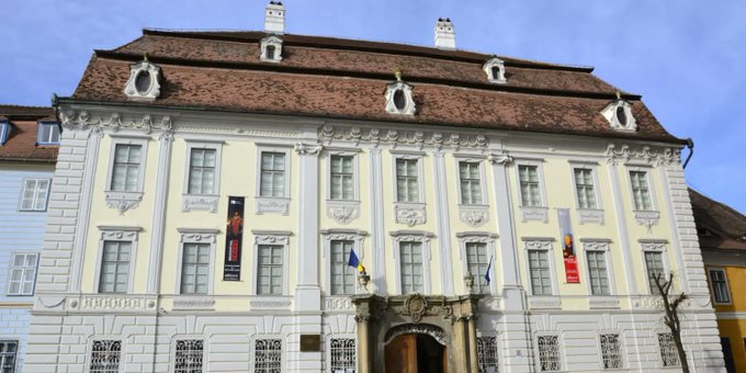 A large baroque-style building with a cream-colored facade, multiple windows, flags hanging from the front, and a brown tiled roof under a blue sky.