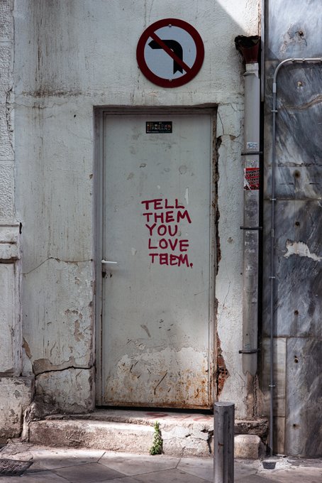 A worn gray door set in a cracked concrete wall, spray-painted with the red message 'TELL THEM YOU LOVE THEM,' located below a circular 'no dogs' sign.