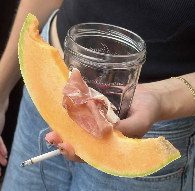 A slice of cantaloupe with prosciutto and a glass of red wine is held by a woman in jeans, with a lit cigarette balanced on the cantaloupe, fitting the theme of "The 28-year-old Italian girl lunch (2026)". The glass appears to be a "Le Parfait" jar.