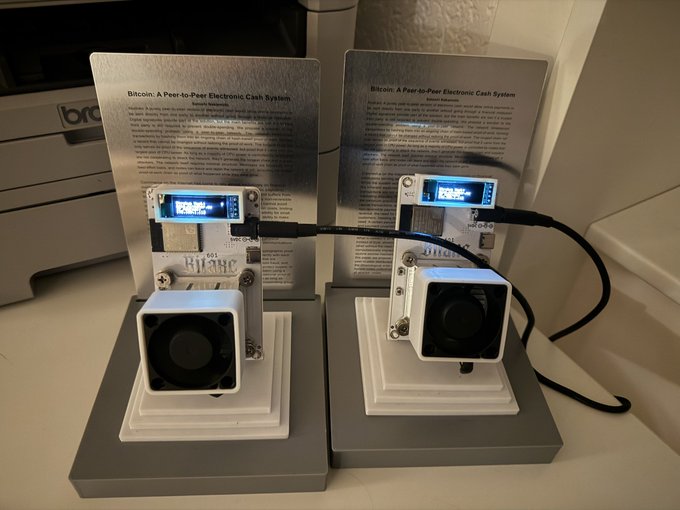 Two Bitcoin mining devices displayed on stands in front of informational placards titled "Bitcoin: A Peer-to-Peer Electronic Cash System," featuring small screens and cooling fans, suggesting the wife encountered these unfamiliar gadgets while unboxing them in the user's absence.