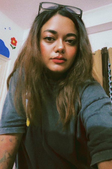 A selfie of a young woman with long dark hair wearing a dark gray top, with sunglasses perched on her head, taken indoors with a flower decoration visible on the wall.