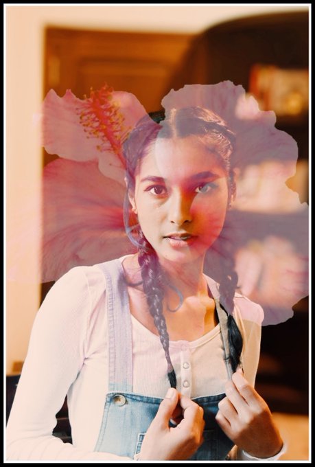 A young woman with braided hair and overalls gazes directly at the viewer, overlaid with the translucent pink silhouette of a hibiscus bloom to create a soft double-exposure effect that visually echoes the artwork's theme of fading presence and delicate beauty.