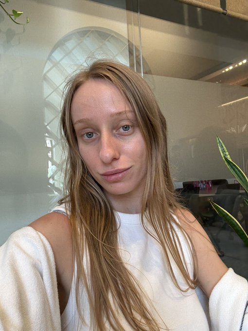 A selfie of a young woman with long blonde hair wearing a white top, taken indoors with plants in the background.