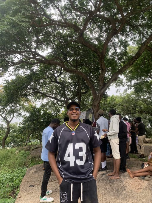 A young man wearing a black jersey with the number '43' poses for a photo outdoors under a large tree, with other people gathered in the background on a rocky outcrop.