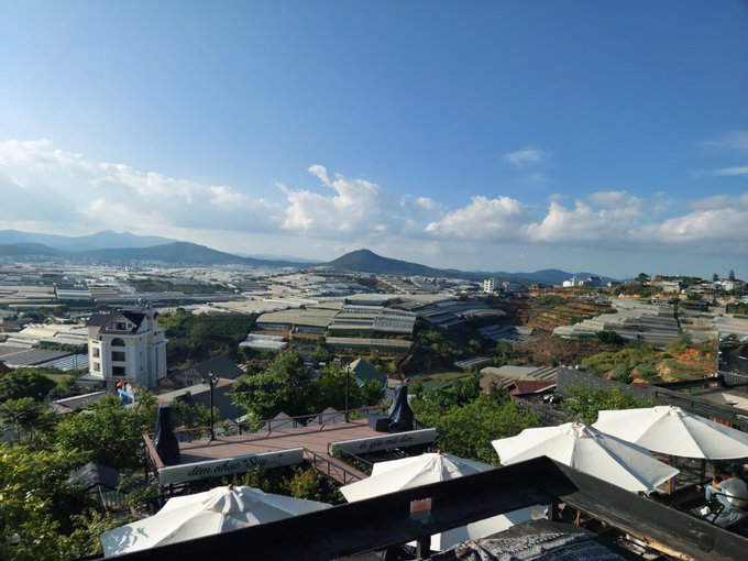 The image shows a cityscape with buildings of various sizes under a blue sky dotted with clouds. The scene appears to be taken from an elevated position, possibly from a rooftop or balcony. In the foreground, there are several white umbrellas providing shade for people below. The image also contains text that reads "gMorning The gap between blockchain infrastructure and real world adoption is getting smaller. With the satUSD Payment SDK, developers can integrate cross chain payments directly into apps, game and merchant tools built on @RiverdotInc , making payments more seamless for users."