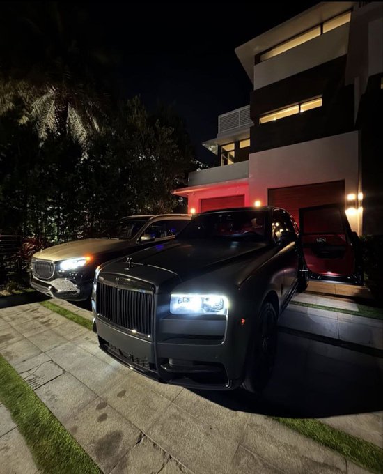 A Rolls-Royce Ghost and a BMW are parked at night in the driveway of a modern, multi-story home with illuminated windows. The caption "Can I get a GN?" plays on the Rolls-Royce Ghost ("G") and the vehicle's nighttime lighting, suggesting a request to show off the luxury car.