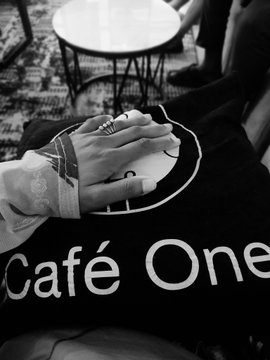 A black and white photograph shows a tattooed hand resting on a lap, with rings visible on the fingers, near a person wearing a shirt reading 'Café One' with a small table in the background.