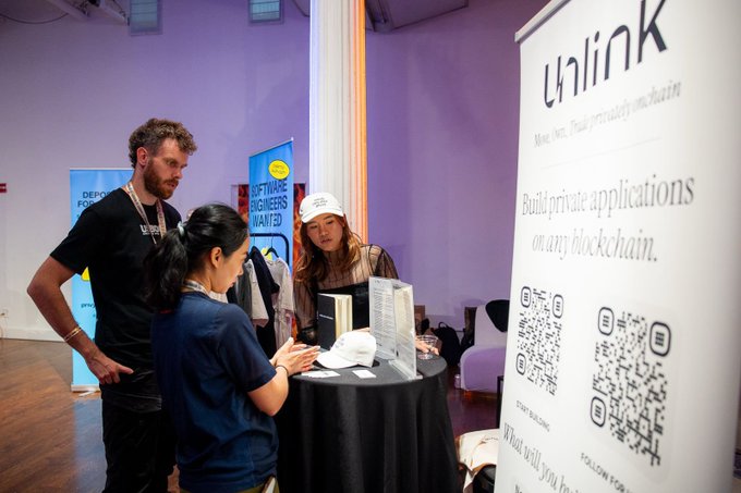 The image shows a group of people gathered around a table with a sign that says "Unlink" above them. The people are engaged in conversation, possibly discussing the benefits of using Unlink for private applications or payments. One person appears to be reading something from a book on the table while others look at it. There is also an open laptop and a keyboard visible on the table.
