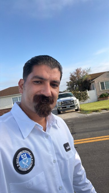 A selfie of a man with a mustache and goatee wearing a white button-up shirt featuring a 'Proud Member of the 924- Photojournalism Education' patch, standing on a suburban street.