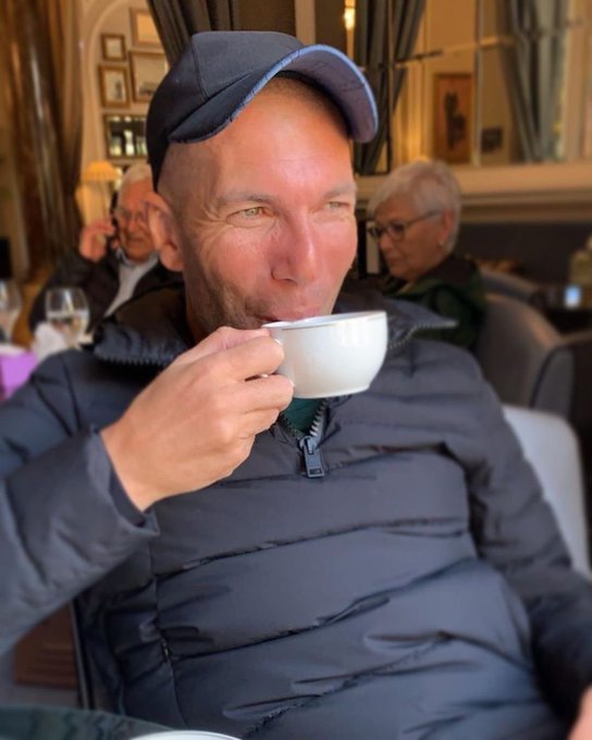 A smiling man wearing a blue cap and a dark puffy jacket sips from a white coffee cup while seated in what appears to be a restaurant or lounge.