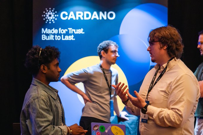 Two developers engage in conversation at the Cardano Origins Hackathon, with a large backdrop displaying the Cardano logo and the slogan "Made for Trust. Built to Last." behind them, capturing the collaborative energy as participants prepare to tackle their 36-hour build challenge with x402.