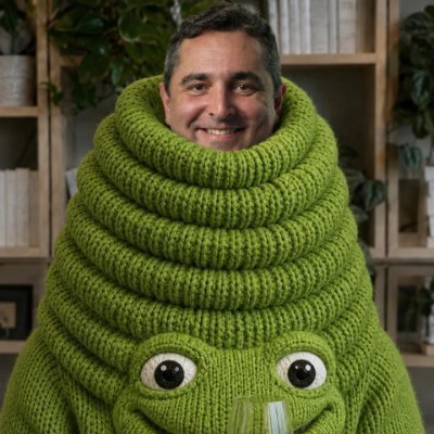 A smiling man wearing a bright green knitted frog/cartoon hoodie poses in front of wooden shelves. The whimsical, meme-style image ties into the playful $TOAD token giveaway request, using the frog-like aesthetic to match the amphibian-themed crypto promotion.