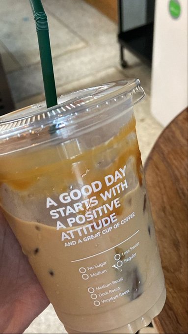 A hand holds a plastic cup of iced coffee with a green straw; the cup is printed with "A GOOD DAY STARTS WITH A POSITIVE ATTITUDE AND A GREAT CUP OF COFFEE" and roast options.