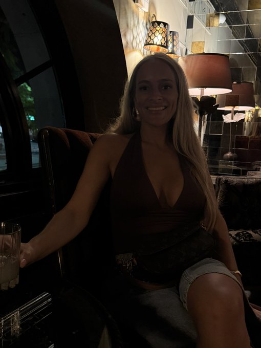 A smiling woman with long blonde hair wearing a dark halter top sits in a dimly lit lounge or bar, holding a drink.